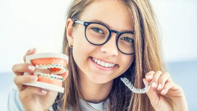 A teenage girl holding up a tooth model with metal braces in one hand and an Invisalign retainer in the other.